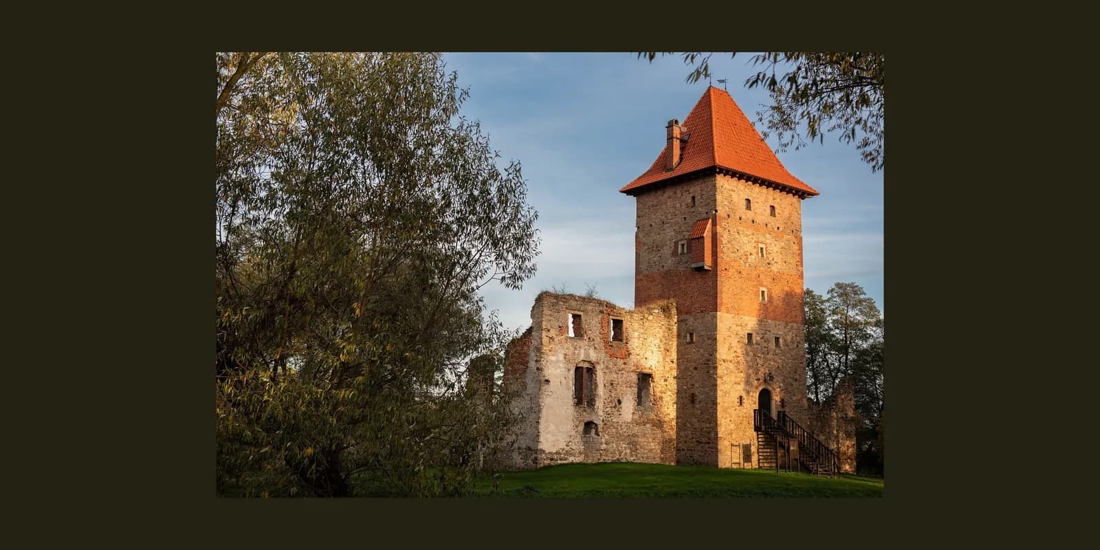 A historic stone tower with a red-tiled roof and partial ruins, surrounded by trees under a blue sky.