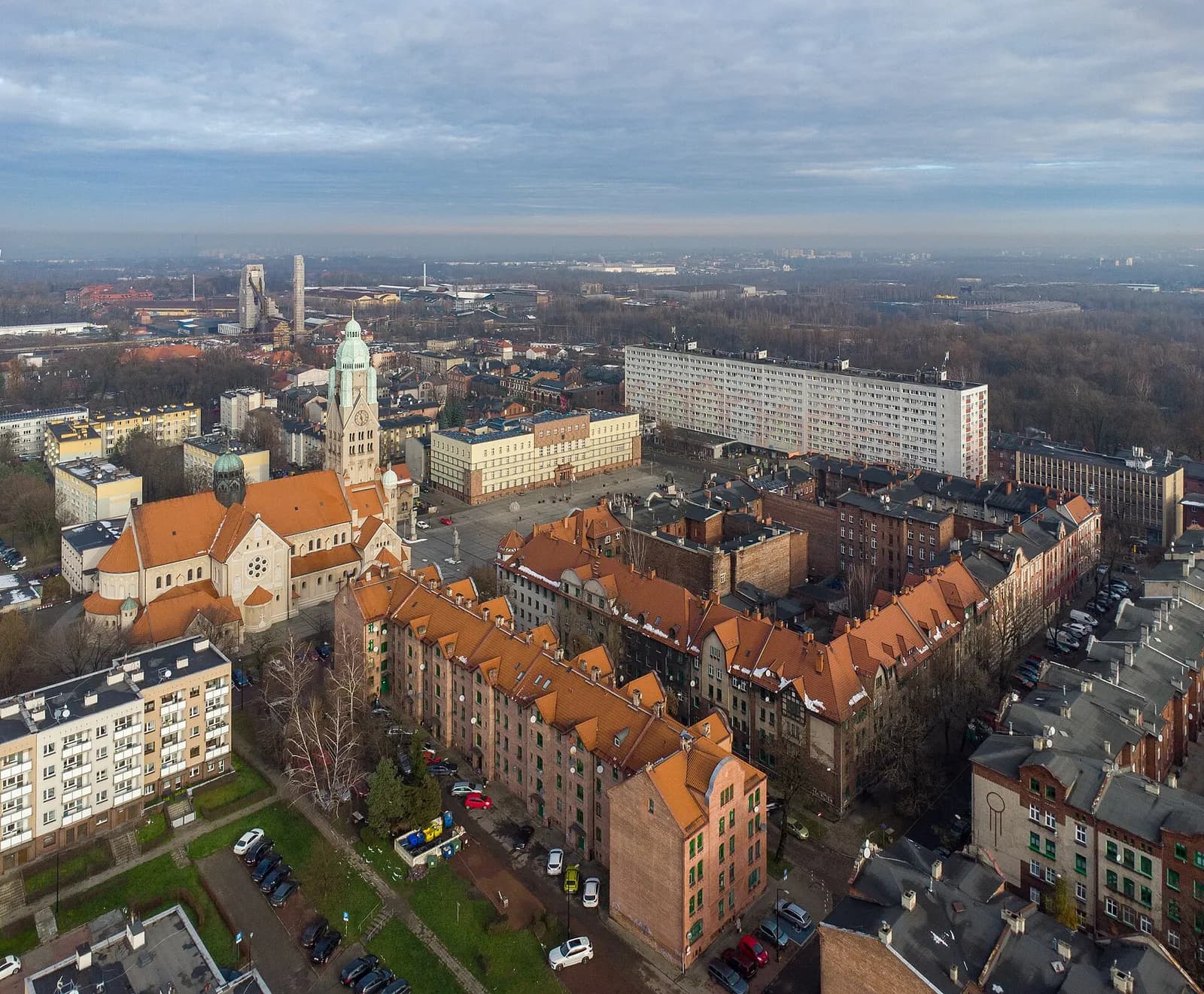 An aerial view of a town, showing a large brick church with a green-domed tower, surrounded by various residential buildings with red-tiled roofs, and a distant cityscape with industrial structures under a cloudy sky.